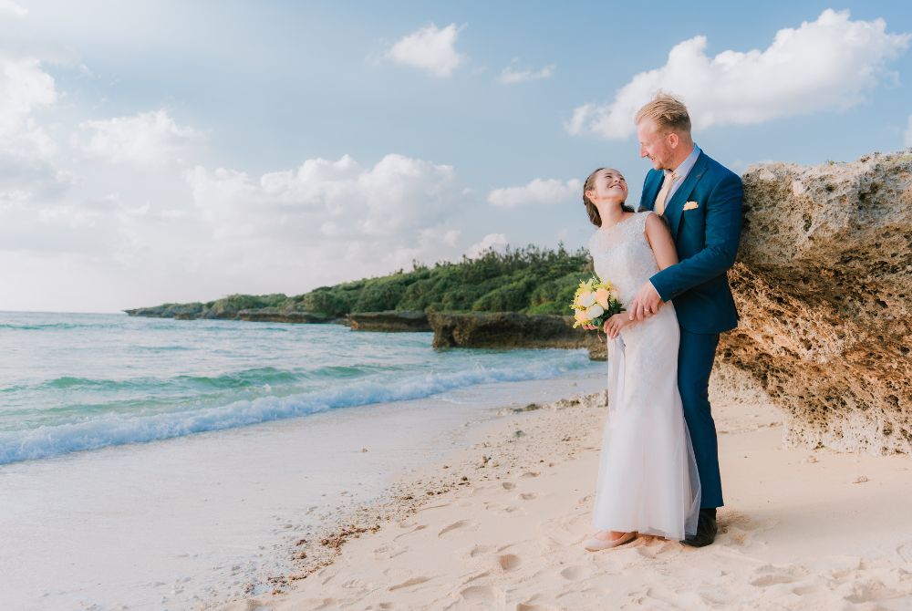 青いタキシードの新郎とホワイトドレスの新婦がビーチで岩場をバックに互いを見つめて撮影
